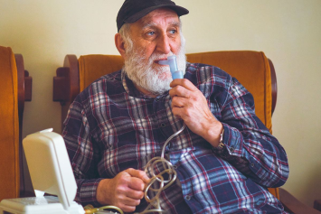 A person sits in a cushioned chair and holds a plastic breathing device to their mouth.