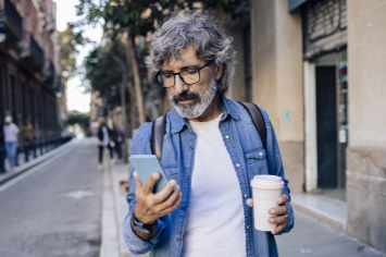 A person looks at their phone on a sidewalk holding a coffee.