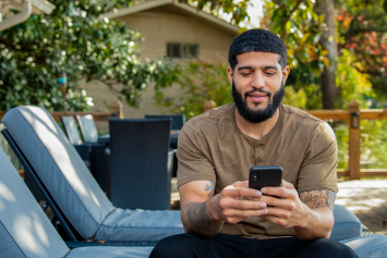 A Veteran sits on a lounge chair outside while looking at their phone.