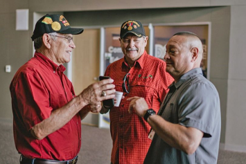 Three people stand in a group talking. Two are wearing hats embroidered with Veteran insignia, and one shows the others something on his phone.  