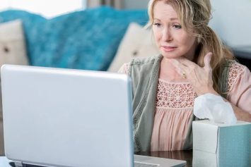 A person holds one hand to their throat while sitting at home with a box of tissues to the side and a laptop in front of them.